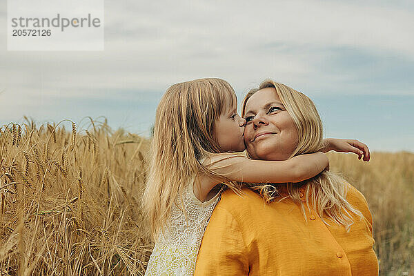 Blond hair girl kissing mother in wheat field under sky