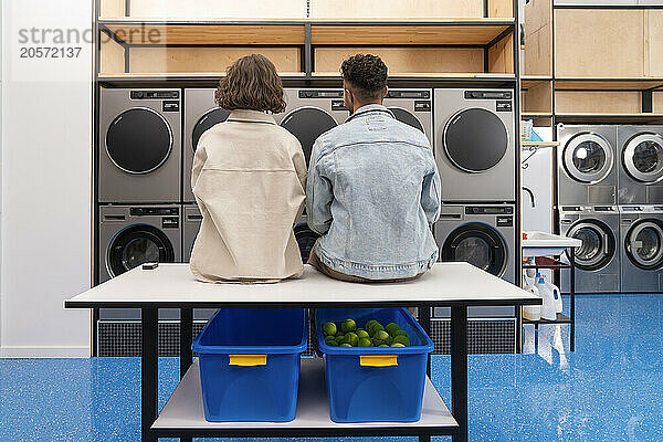 Young couple sitting on table in front of washing machines at laundromat