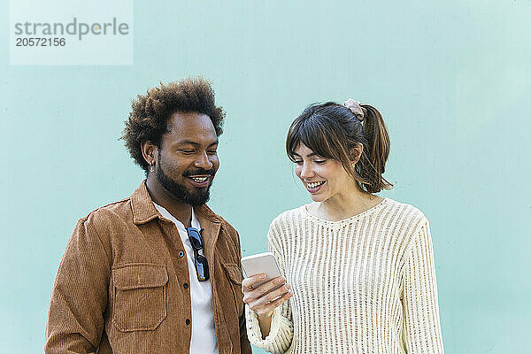 Happy woman sharing smart phone with boyfriend in front of blue wall