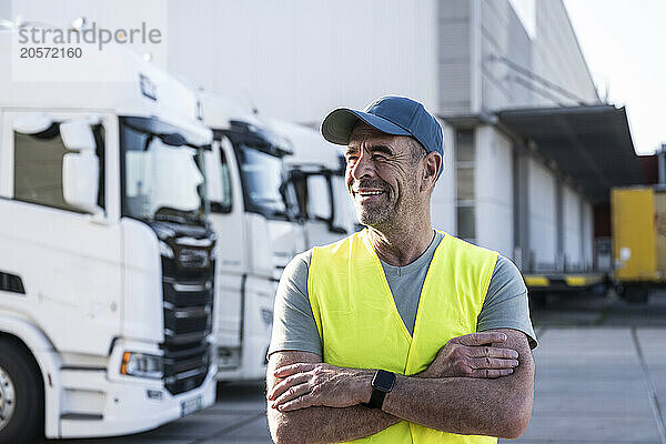 Smiling mature dock worker standing with arms crossed looking away outside warehouse