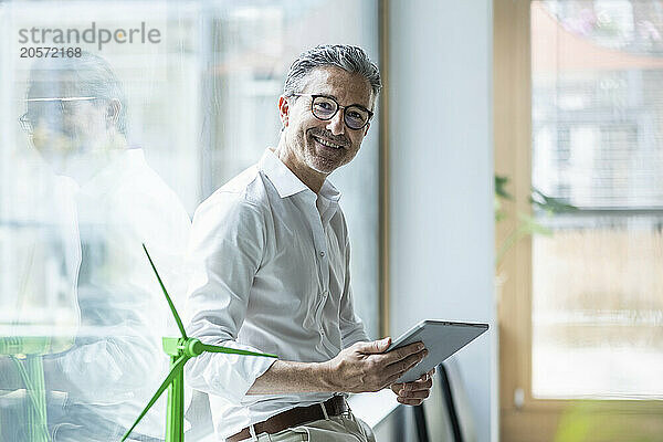 Smiling mature businessman holding tablet PC leaning on window by wind turbine model