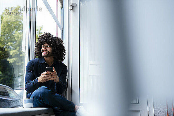 Smiling businessman with smart phone sitting on window sill looking out at office