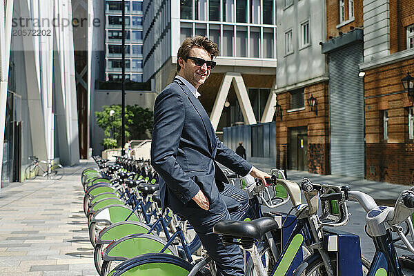 Happy businessman wearing sunglasses and renting bicycle near buildings on sunny day