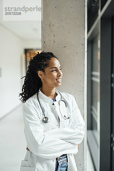 Smiling young female doctor with arms crossed leaning on column looking out through window at hospital