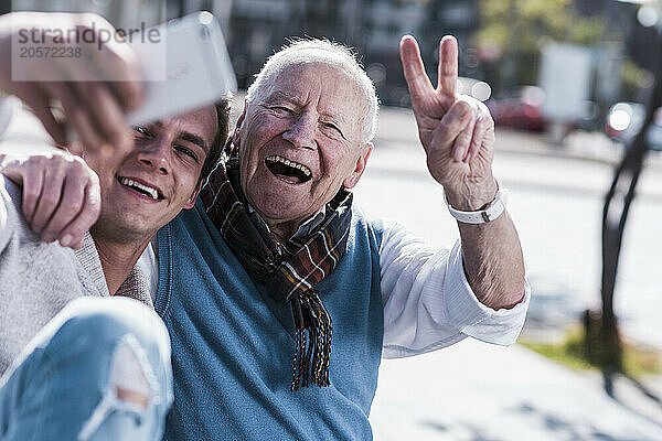 Happy senior man gesturing peace sign with grandson taking selfie through smart phone