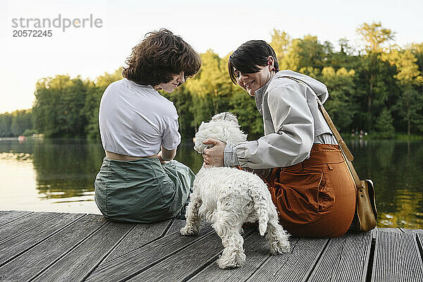 Mother and daughter with white dog sitting on pier at river