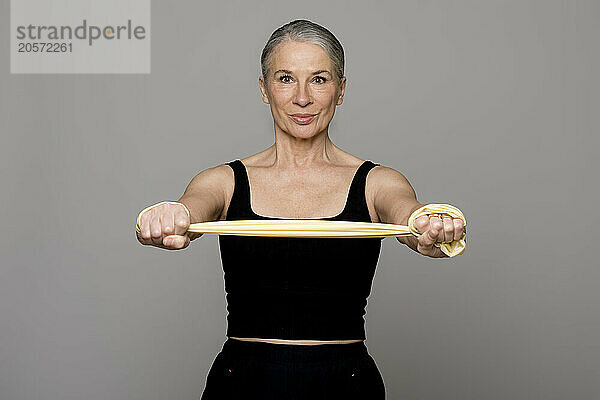 Confident senior woman in black sports clothing exercising with resistance band against gray background