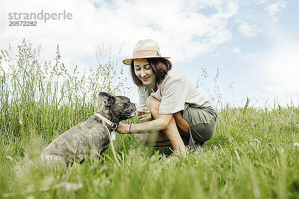 Smiling woman kneeling and petting french bulldog in field