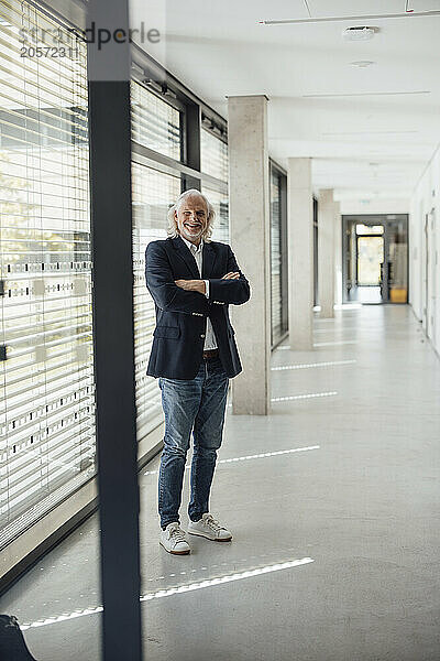 Happy senior businessman standing with arms crossed in office corridor