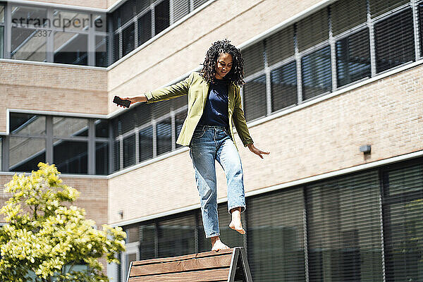 Young businesswoman balancing with arms outstretched on bench at office park