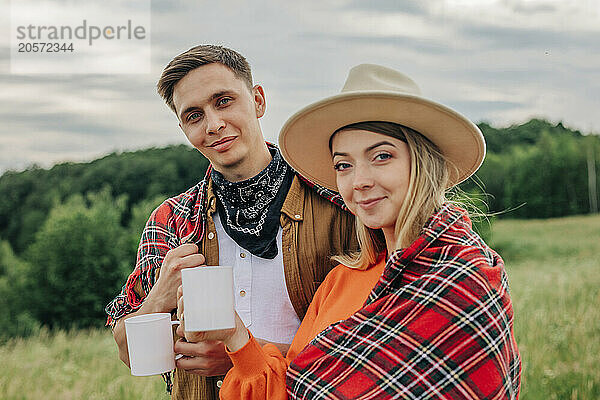 Smiling young couple in plaid shawl holding tea mug on meadow