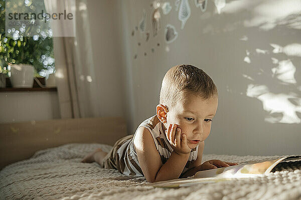 Cute boy lying on bed reading picture book at home