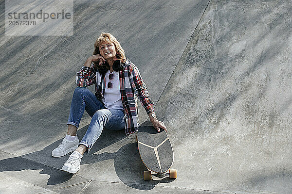 Smiling senior woman sitting with skateboard on sports ramp in park