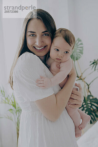 Smiling woman holding baby boy and standing at home