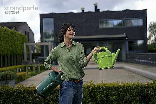 Smiling businesswoman holding watering cans in front of house