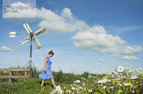 Mature woman walking near flowers and windmill under clouds in field