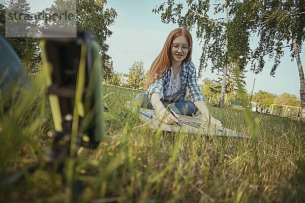 Teenage influencer making protest banner and vlogging through smart phone sitting on grass at park