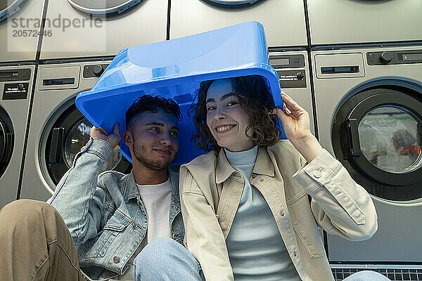 Happy young couple with blue basket over heads sitting in laundromat