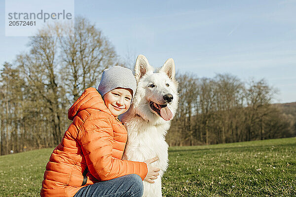 Smiling boy in warm clothes next to White Swiss Shepherd Dog on grass in meadow