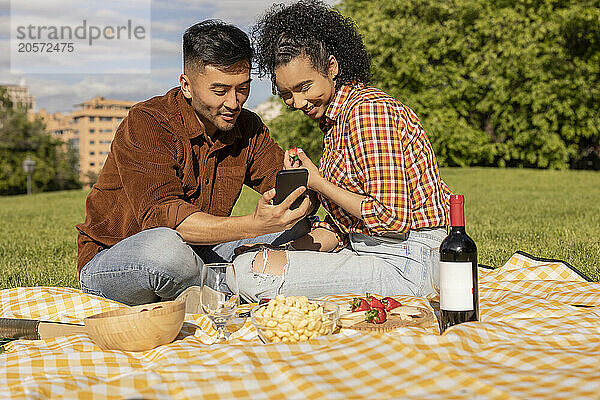 Happy young couple using smart phone and sitting on picnic blanket at park