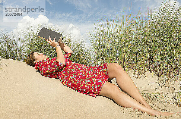 Woman reading book and lying on sand near plants under cloudy sky