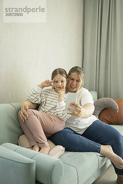 Mother and daughter taking selfie through smart phone sitting on sofa at home