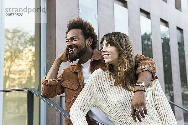 Smiling multiracial couple leaning on railing and looking away