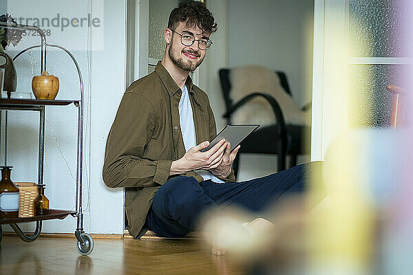 Smiling young handsome man with tablet PC sitting at doorway in living room