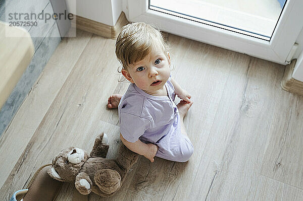 Cute baby girl sitting by teddy bear on floor at home