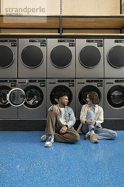 Smiling young couple sitting together leaning on washing machines in laundromat