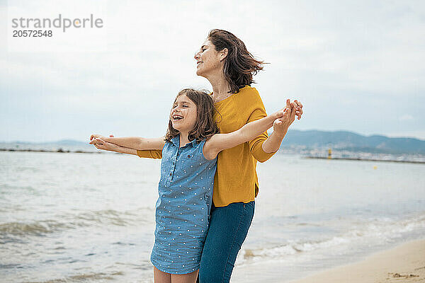 Happy mother and daughter with arms outstretched standing at beach