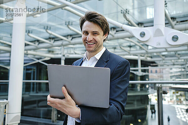 Happy businessman holding laptop and standing near railing