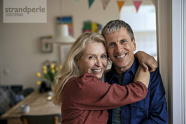 Smiling retired senior woman with arm around man leaning on wall at home