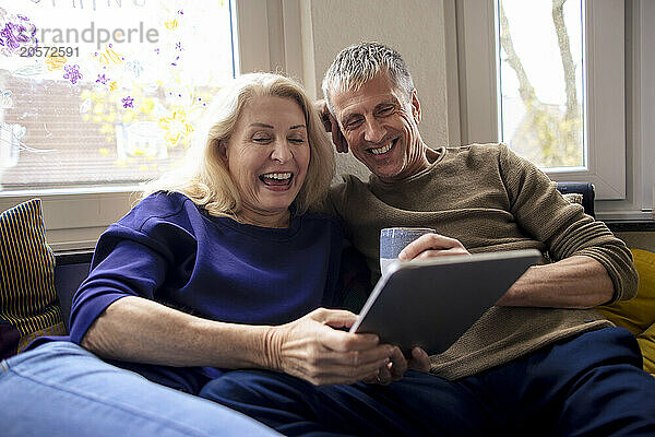 Cheerful senior couple sitting with tablet PC and coffee cup on sofa at home