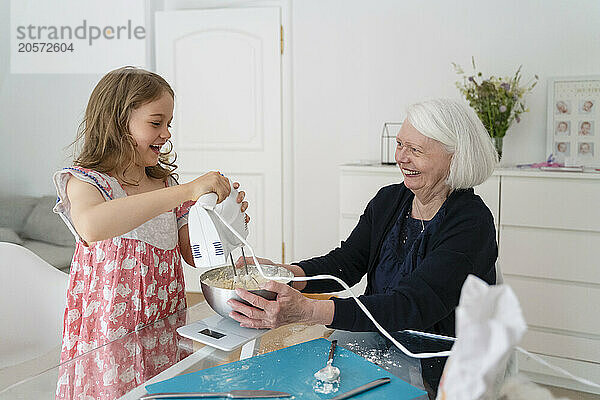 Happy girl mixing dough with blender by grandmother at home