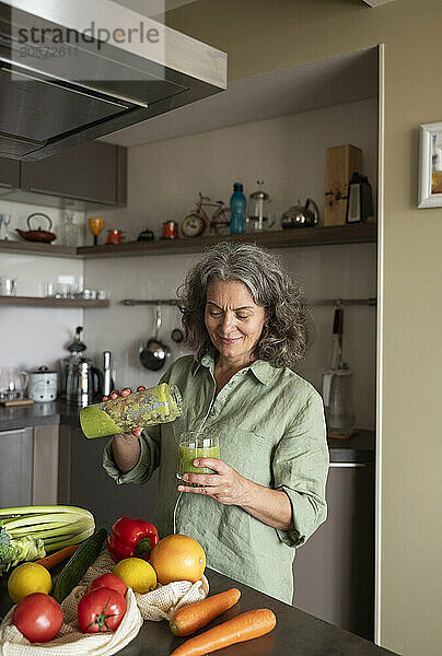 Mature woman poring detox smoothie in glass at kitchen