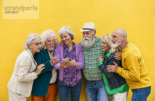 Happy senior woman sharing smart phone with friends in front of wall