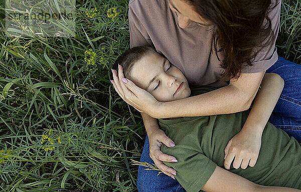 The boy lies with his eyes closed on his mother's lap on the grass.