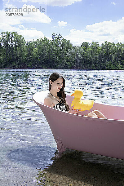 Smiling girl holding yellow rubber duck and sitting inside pink bathtub in lake