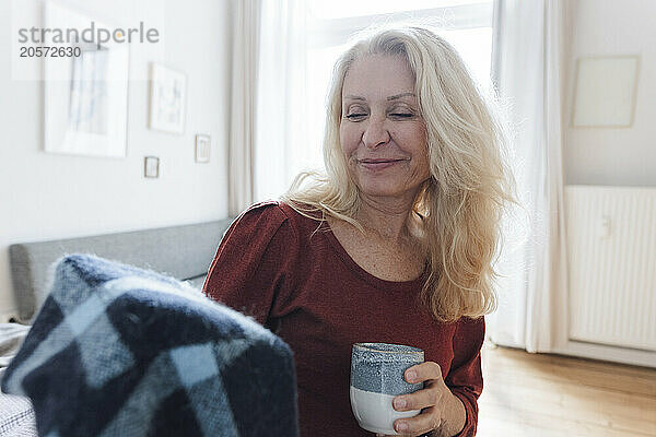 Smiling blond woman with coffee cup looking at blanket in living room