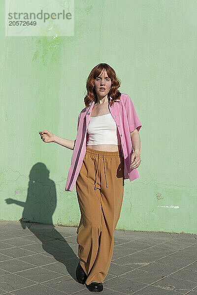 Confident redhead woman dancing on footpath in front of mint green wall