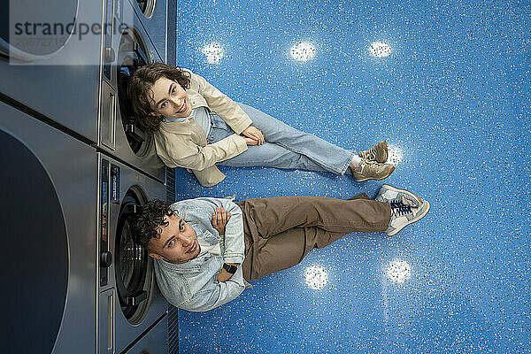 Smiling young couple leaning on washing machines sitting in laundromat