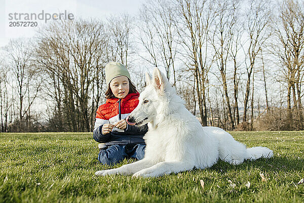 Boy sitting next to White Swiss Shepherd Dog on grass in meadow