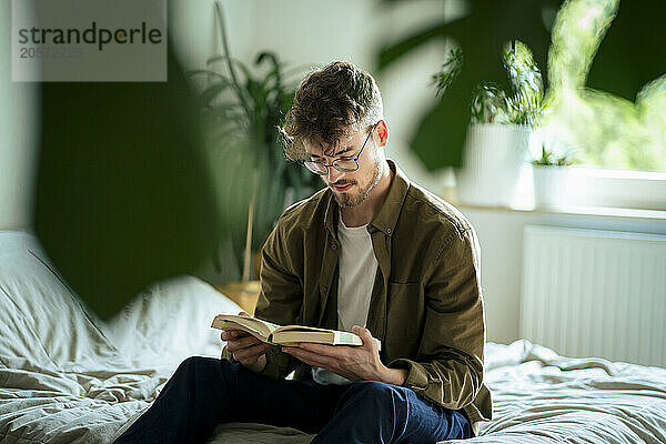 Young man reading book sitting on bed at home