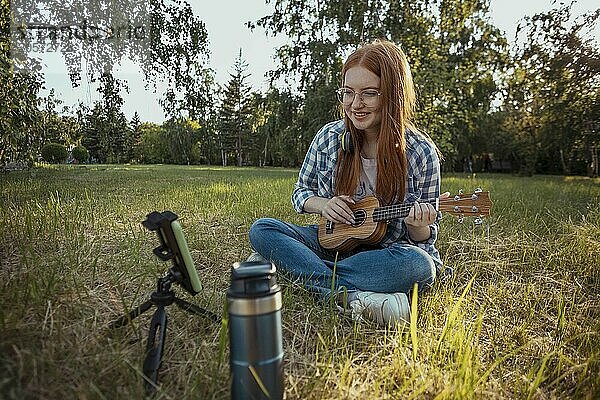 Teenage influencer playing ukulele and vlogging through smart phone at public park