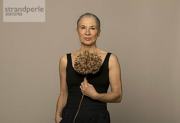 Woman with hands in pockets holding withered flower against brown background