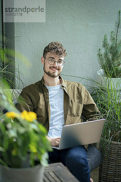 Smiling handsome young man sitting with laptop in balcony