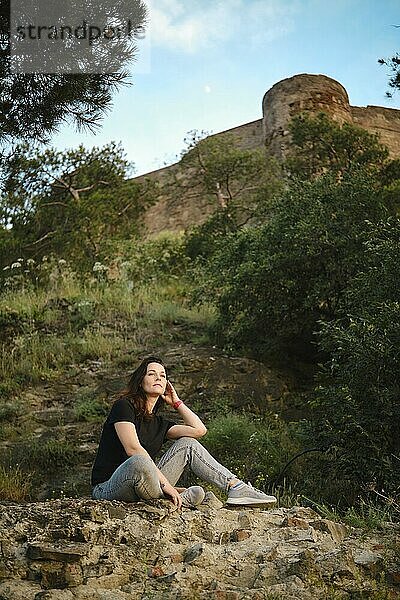 Dreamy woman sitting amidst the rocky terrain and lush greenery  with a hillside and ancient fortress on background