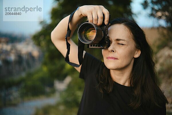 A woman photographer climbed a high hill and photographs the city below illuminated by the rays of the setting sun (focus on camera lens)