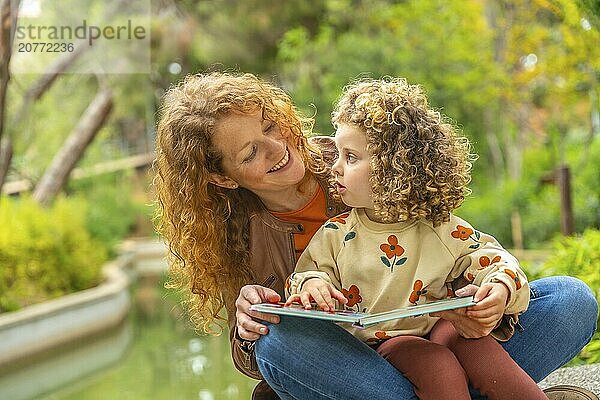 Cute girl and smiling caucasian mother reading a book sitting on a park together